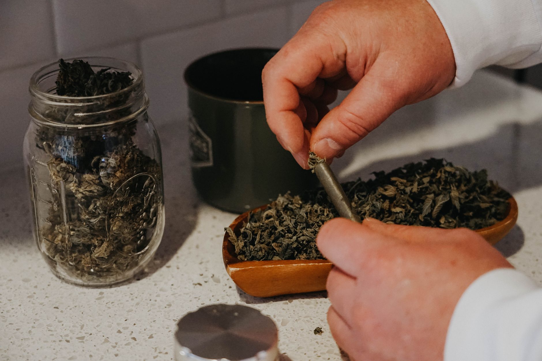 A man skillfully rolling cannabis joint with herbs on a wooden tray and jar nearby.